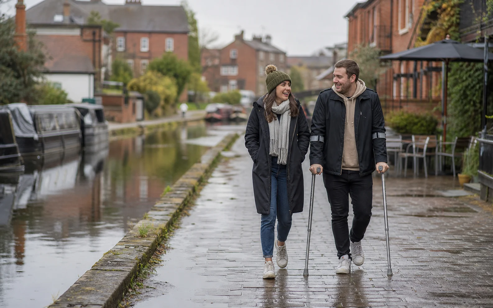 A British couple walking beside a canal after rain, with one partner using forearm crutches